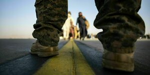 Durch die Beine eines US-Soldaten fotografiert sind afghanische Flüchtende unscharf zu erkennen