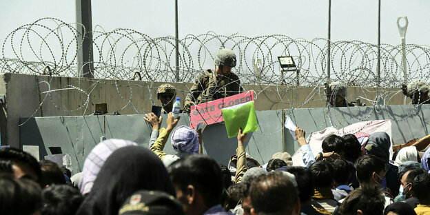 Menschen drängeln sich mit Papieren in der Hand vor der Flughafenmauer mit Stacheldraht, die von US Soldaten bewacht wird