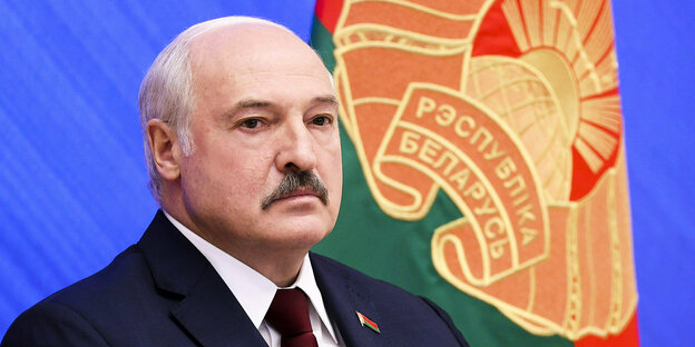 Portrait von Lukaschenko mit ernstem Gesichtsausdruck vor blauem Hintergrund mit Flagge