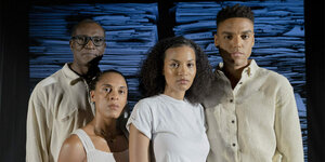 Gruppenbild: Die Schauspieler:innen des Stücks "Courageous! Furchtlos!", Aloysius Itoka, Maya Alban-Zapata, Amina Eisner und Lamin Leroy Gibba, stehen von einem Blau-Schwarzen Hintergrund