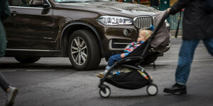 Ein Kinderwagen wird vor einem SUV über die Straße geschoben