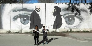Zwei Kinder spielen vor einer Wand mit Stacheldrahtzaun. Auf der wand ist ein Graffity von dem Handschlag zwischen Khalizad und Mullah Abdul Ghani Bardar