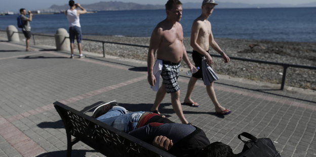 Touristen auf der Insel Kos. Auf einer Bank ruht sich ein syrischer Flüchtling aus