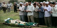 Männer mit traditionellen Kopfbedecken beten vor einer Trage, auf der die Tote liegt