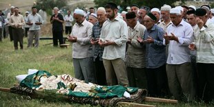 Männer mit traditionellen Kopfbedecken beten vor einer Trage, auf der die Tote liegt