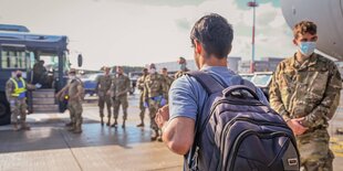 Ein Mann mit einem rucksack geht an einem Flughafen an Soldaten vorbei. Im Hintergrund ein Bus