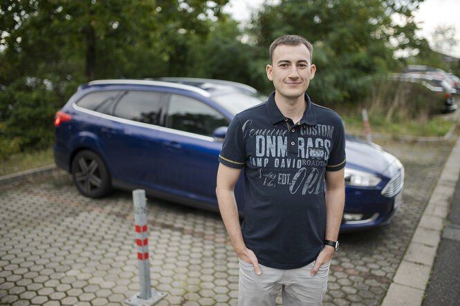 Eine Personmit kurzen Haaren und dunkem Poloshirt steht auf einem Parkplatz vor einem blauen Auto