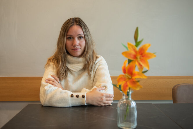 Eine Person mit langen blonden Haaren und einem weißen Rollkragenpullover sitzt an einem Tisch und blickt in die Kamera