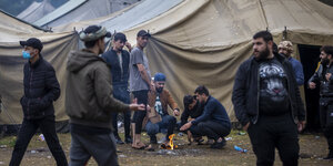 Eine kleine Gruppe von Männern bereitet in einem neu errichteten Flüchtlingslager auf dem Truppenübungsplatz Rudninkai ein Feuer, wohl zur Essenszubereitung, vor. Hinter ihnen sind mehrere große beige Zelte zu sehen