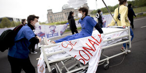 Zwei Mediziner:innen protestieren vor dem Berliner Reichstagsgebäude auf einem Krankenbett für bessere Arbeitsbedingungen.