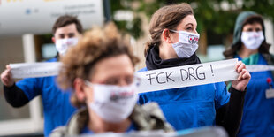 Menschen tragen Masken und blaue Kittel. Eine Person hält einen Spruch hoch: "Fck DRG" steht darauf.