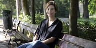 Hanna Schoeville sitzt mit einer schwarzen Bluse und Jeans bekleidet auf einer Parkbank und lächelt