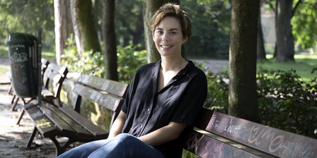 Hanna Schoeville sitzt mit einer schwarzen Bluse und Jeans bekleidet auf einer Parkbank und lächelt