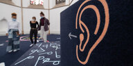 Ein Ausstellungsraum mit einer Wand, auf dem ein Ohr und ein Pfeil aufgezeichnet sind. Auf dem Boden sind die Buchstaben "p" und "q" aufgemalt.