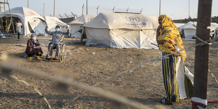 ein mann mit einem Rollstuhl zeigt mit dem Daumen nahc unten, neben ihm kauert eine Frau, dahinter weiße Zelte des UNHCR und Stacheldraht, eine Frau mit gelbem Kopftuch betritt von rechts die Szene