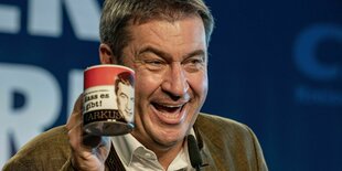 Markus Söder hält eine Markus Söder Tasse in der Hand und strahlt