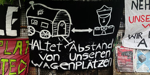 Ein schwarzes Banner an einem Zaun, darauf ein Polizist und ein Wohnwagen in weißer Farbe aufgemalt