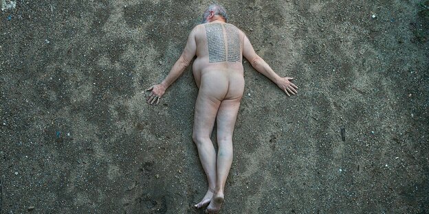 Ein nackter Mann liegt bäuchlings auf dem Boden, auf seinem Rücken sind Sätze geschrieben.