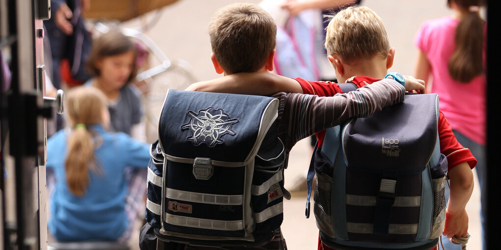 Zwei Jungen mit großen Schulranzen auf dem Rücken legen einander die Arme um die Schultern.