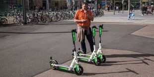 Ein Mann steht vor einem Bahnhof mit einem Smartphone und E-Rollern