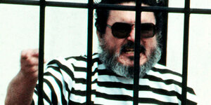 Der Guerilla-Anführer 1992 nach seiner Festnahme hinter Gittern in einem schwarz-weißen Sträflingshemd und Sonnenbrille. Er reckt die Faust und schreit