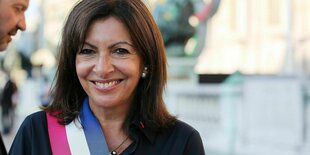 Anne Hidalgo lächelt in die Kamera, sie trägt offenes Haar und eine Schärpe in den französischen Nationalfarben über dem dunklen Oberteil