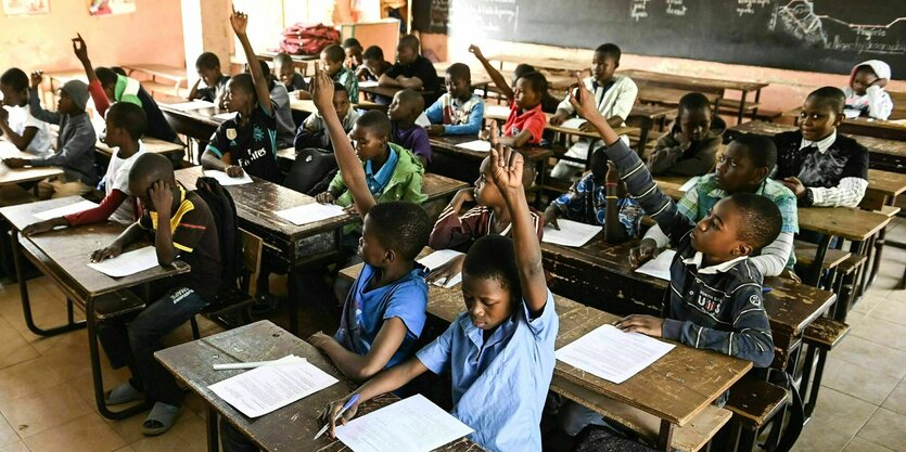 Kinder sitzen an Holzbänken in einer Schule und melden sich vor der Lehrerin