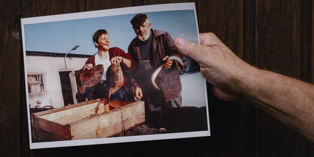 Eine Hnd hält ein älteres Foto, auf dem Angela Merkel mit zwei Fischen in der Hand und ein Fischer zu sehen sind