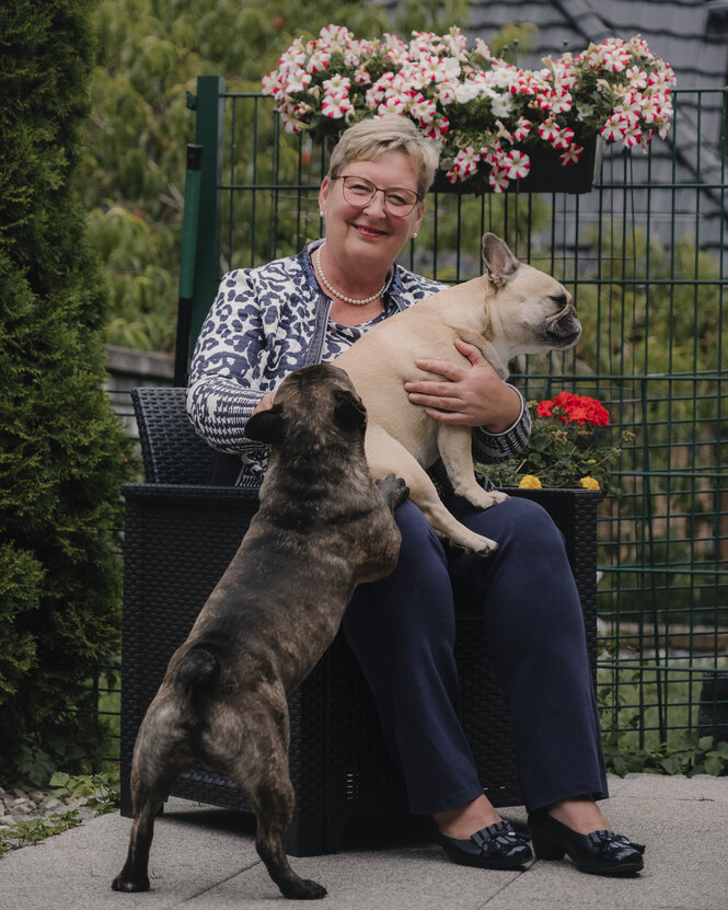 Eine Frau sitzt auf einem Stuhl, eine Bulldogge auf ihrem Schoß, eine springt an ihr hoch