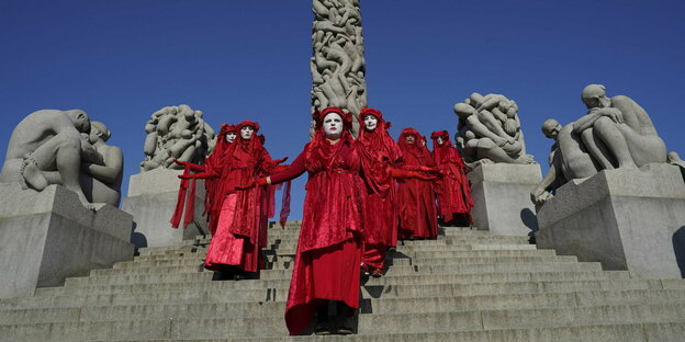 rot gewandete Klimaaktivisten mit weißen Masken prozessieren zwischen Steinskulpturen