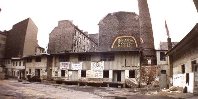 Ein trister Hinterhof mit Berliner Altbauten, einem Schornstein und einem Haus, das mit einem Rgeenbogen und dem Wort "Instandbesetzt" bemalt ist, 1981.