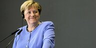 Angela Merkel steht an einem Rednerpult und lächelt verschmitzt