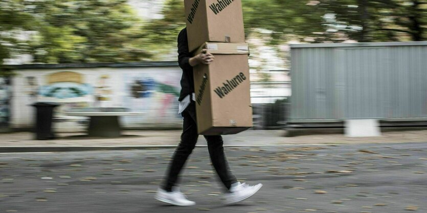 Ein Wahlhelfer trägt zwei Wahlurnen aus Karton, die ihn selbst überragen