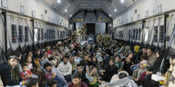 Viele Menschen sitzen im transportraum eines Militär-Flugzeugs, im Hintergrund Soldaten