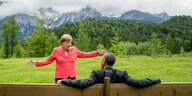 Barack Obama sitzt mit dem Rücken zu den Betrachter:innen auf einer Bank, die auf einer Wiese steht. Angela Merkel steht ihm gegenüber und breitet die Arme aus