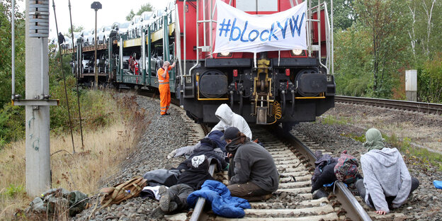 drei Blockierer sitzen auf Gleisen vor enem Güterzug, der mit Autos beladen ist