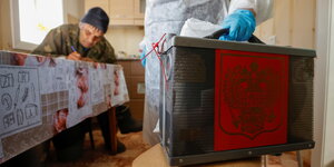 Ein Mann sitzt in seiner Küche und liest einen Stimmzettel während ein wahlhelfer mit der Wahlurne daneben steht.