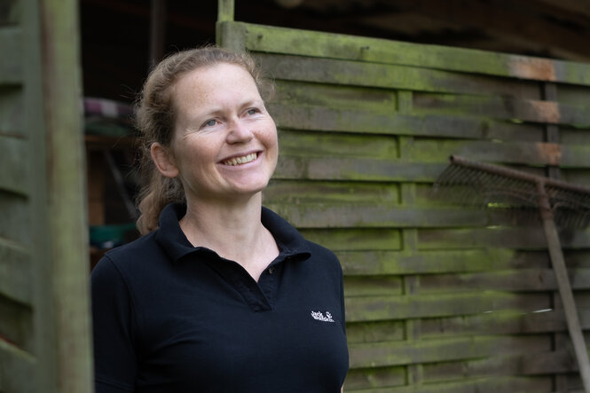 Ute Volz, eine Frau mittleren Alters mit Zopf,vor einem hohen Gartenzaun