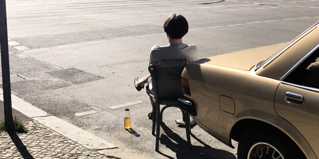 Ein Mann sitzt mit einem Glas Bier, das auf der Straße steht, vor einem geparkten Auto