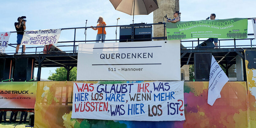 Eine Brücke ist voll gehängt mit Plakaten, eine Frau in der Mitte vor einem Mikrofon. Es handelt sich um eine Demonstration der Querdenken -Bewegung