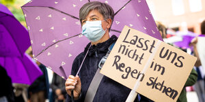Eine Frau hält einen lila Regenschirm und ein Protestplakat mit der Aufschrift "Lasst uns nicht im Regen stehen"