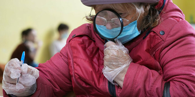 Ältere Frau mit Lupe. Mundschutz und Handschuhen beim Ankreuzen eines Wahlzettels