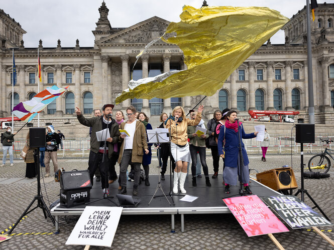 Eine Gruppe von Menscchen schwingt eine goldene Fahne, singt und steht auf einem Podest vor dem Reichstag