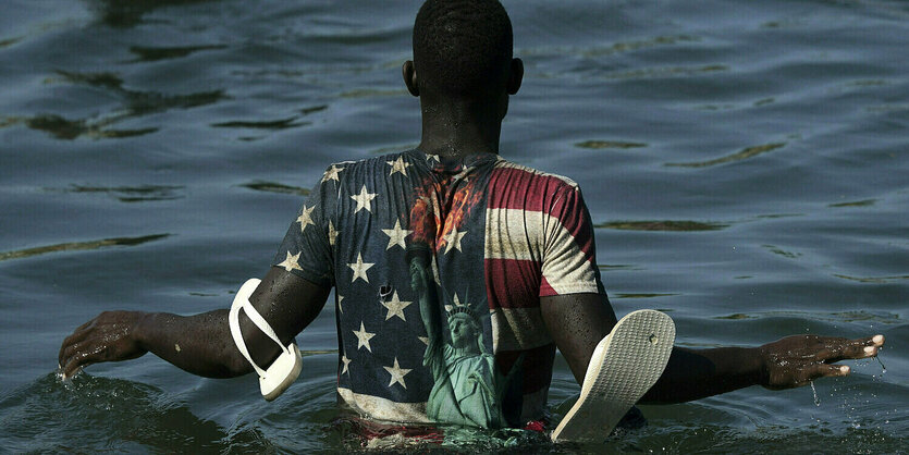 Ein Geflüchteter watet durch den Rio Grande. Er trägt ein T-shirt in den farben der US-Fahne