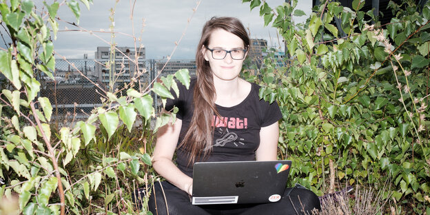 Eine junge Frau mit langen, dunklen Haaren und Brille sitzt mit Laptop zwischen Pflanzen