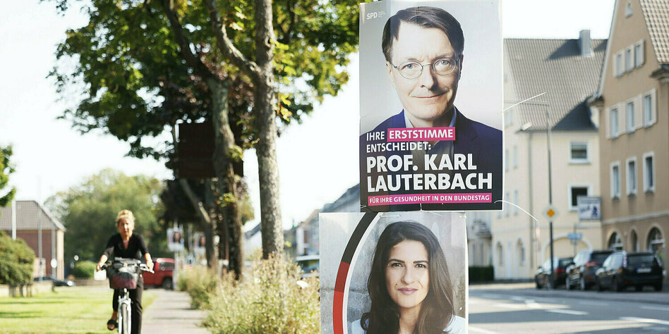 Zwischen einem Fahrradweg und einer bewohnten Straße hängen übereinander zwei Wahlplakate, eins mit Karl Lauterbach, das andere mit Serap Güler