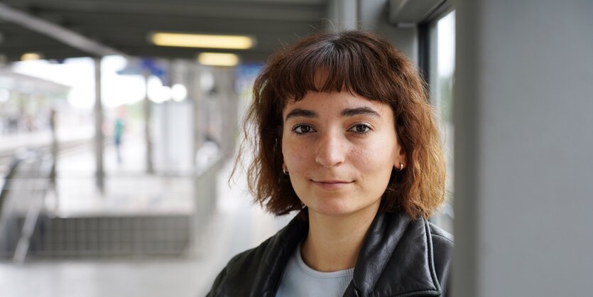 Kathrin Lehmann trägt eine Lederjacke, kurzes Haar und lehnt an einer Säule. Im Hintergrund ist eine Treppe, die nach unten führt.