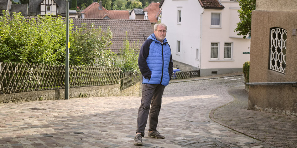 Ein Mann mit blauer Jacke steht mit den Händen in den Jackentaschen auf einer Pflastersteinstraße, im Hitnergrund sieht man eine Hecke und Wohnhäuser
