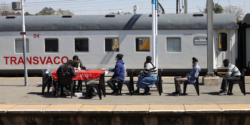 Menscchen sitzen auf Stühlen in Reihen auf einem Bahnsteig vor einem Tisch