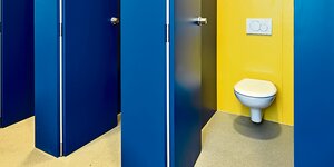 Geöffnete blaue Türen geben einen Blick auf weiße Toiletten vor gelber Wand frei.